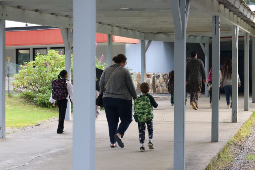 An adult in a gray sweater walks next to a child carrying a green camouflage backpack with their backs facing the camera.