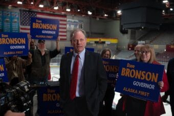 A man in a suit speaking in a hockey stadium surrounded by people holding blue signs with text that says "Dave Bronson, Governor, Alaska First"