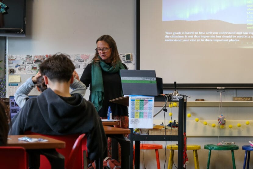 a woman in a black shirt and blue scarf looks down in front of students in a classroom.
