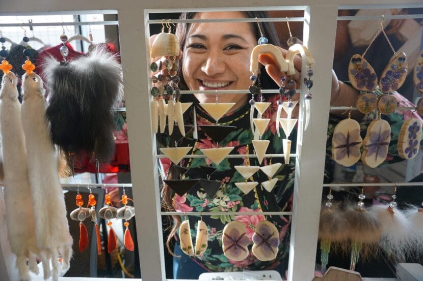 Britt'Nee Brower of Utqiagvik peers through hanging jewelry at her table at the Alaska Federation of Natives convention on Oct. 16, 2025. Brower creates works of art out of a variety of media. Among her skills is carving, sewing, beading, etching, fashion design and poetry. She is among the artists listed in the Alaska Native Arts Directory.