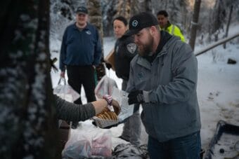 A man in a grey jacket offers a plastic box of cookies to a person out of frame.
