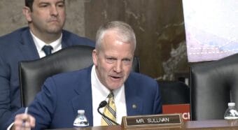 a man in a suit and tie pounds the table in a hearing room. A nameplate identifies him as "Mr. Sullivan."