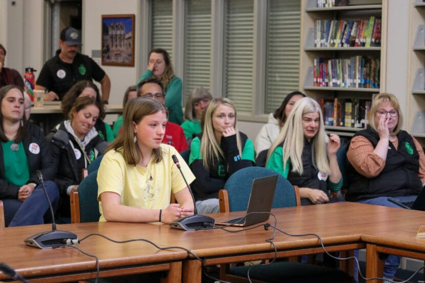Zoe Lessard, dressed in a yellow t-shirt, sits at a wooden table in front of teachers crying during a school board meeting.