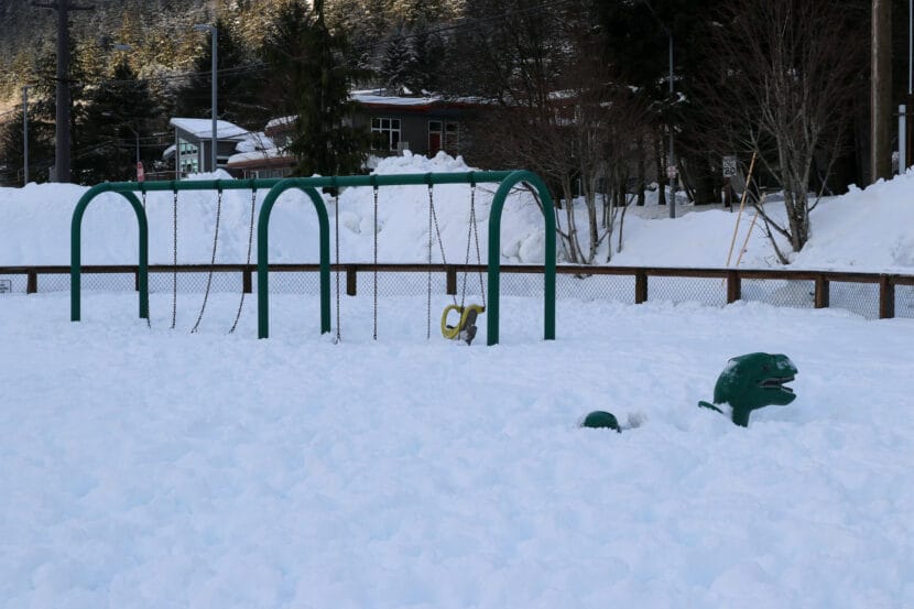 A green dinosaur play structure and a green swing set are covered in several feet of snow.