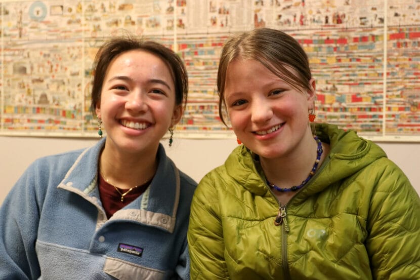 Two students smiled while sitting next to each other. One wears a blue and gray sweatshirt while the other is in a green winter jacket.