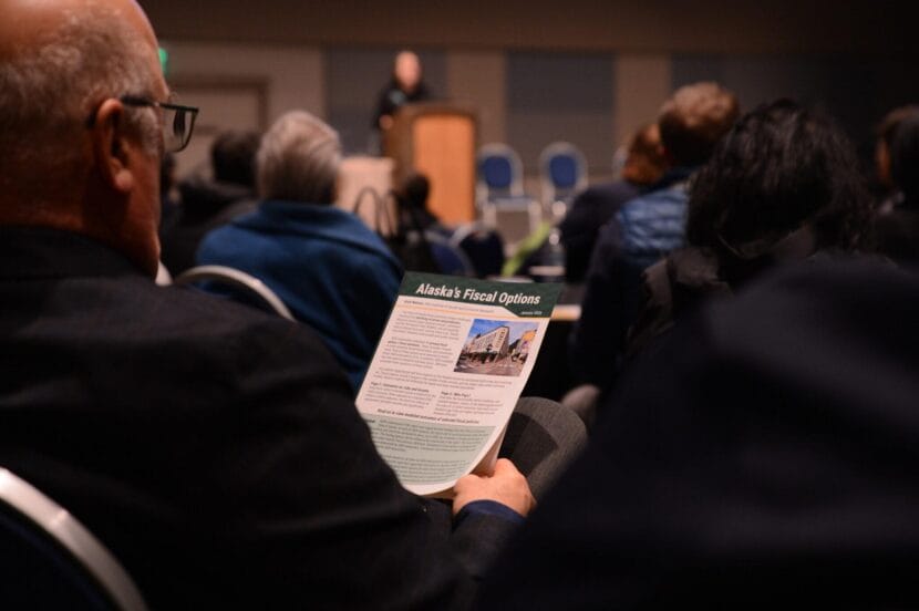 A man sits in the audience of a presentation holding a flyer titled "Alaska's Fiscal Options"