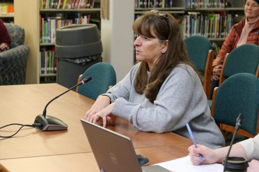 A woman with a gray shirt sits and speaks in front of a small microphone on a wooden table.