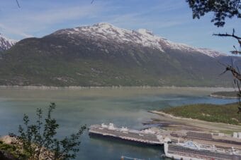 Two cruise ships docked in the foreground of a copper blue body of water, with snow-capped mountains on the opposite shore.
