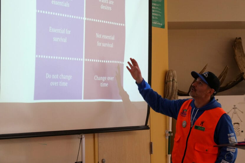 A man in a bright orange vest gestures at a projector screen with his hand. The screen is illuminated with words in boxes.