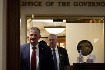 Two men in suits walk down a hallway.